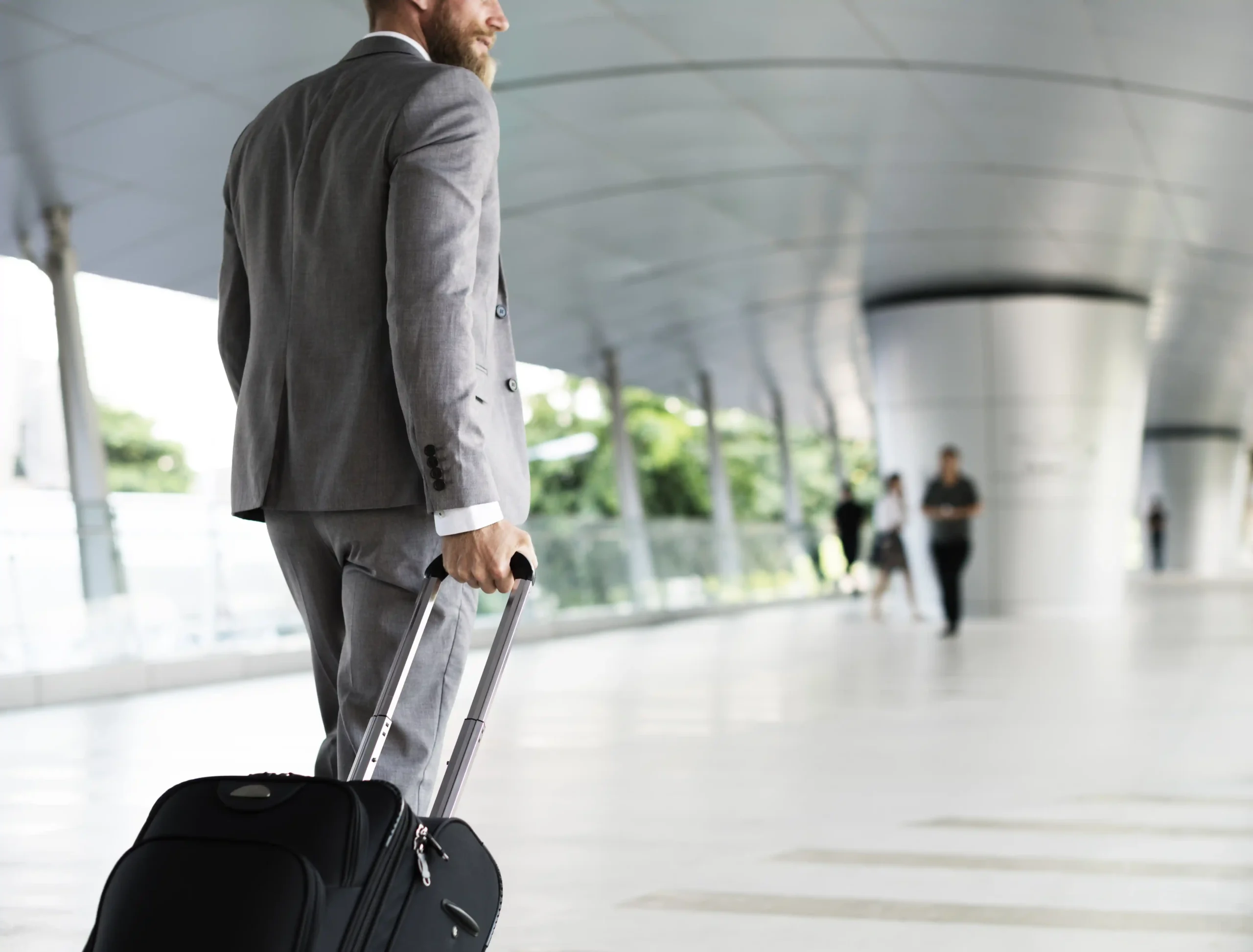 Nell'immagine si vede un uomo con abito elegante che trascina un trolley in un ambiente che ricorda quello di un aeroporto o una stazione. Sullo sfondo, si intravedono altre persone.