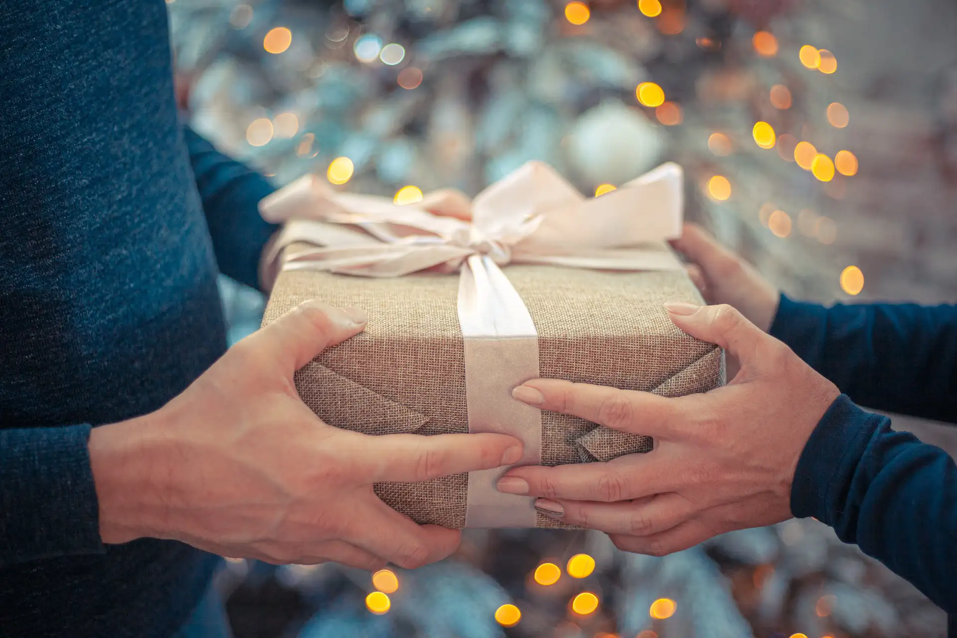 In the picture, the swapping of a gift between two people: we can see two hands holding a present with a white bow. In the background, some blurry lights.