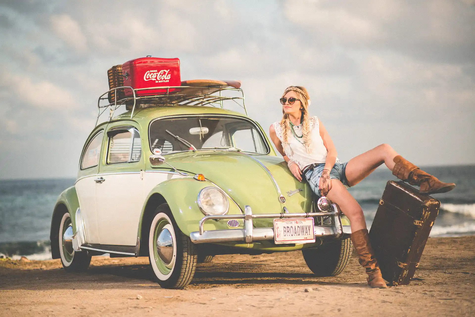 In the picture, a blonde woman with braids leaning on a green car parked on the seaside. She has a leg on a luggage, while some other luggages are piled on the roof of the car.